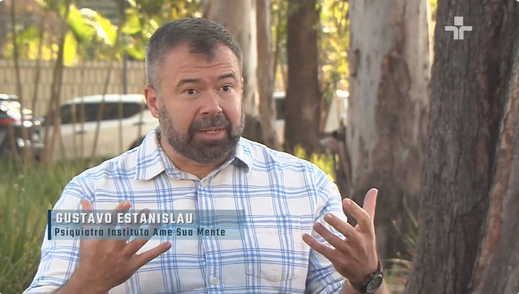 Gustavo Estanislau, psiquiatra da infância e adolescência e especialista do Instituto Ame Sua Mente, está sentado, com camisa xadrez branca e azul e árvores compõem o fundo da cena. Ele destaca os impactos da crise climática na saúde mental infantil.